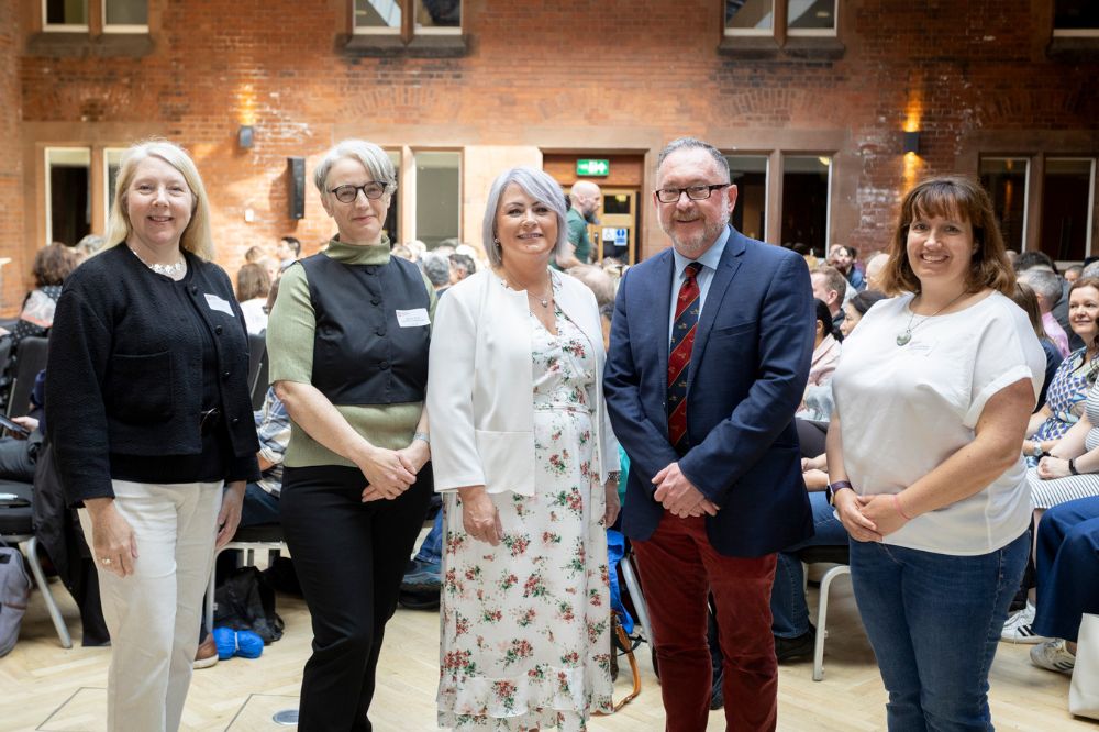 Five individuals (4 female and 1 male), posing standing for a picture in front of a seated audience in a large red brick room
